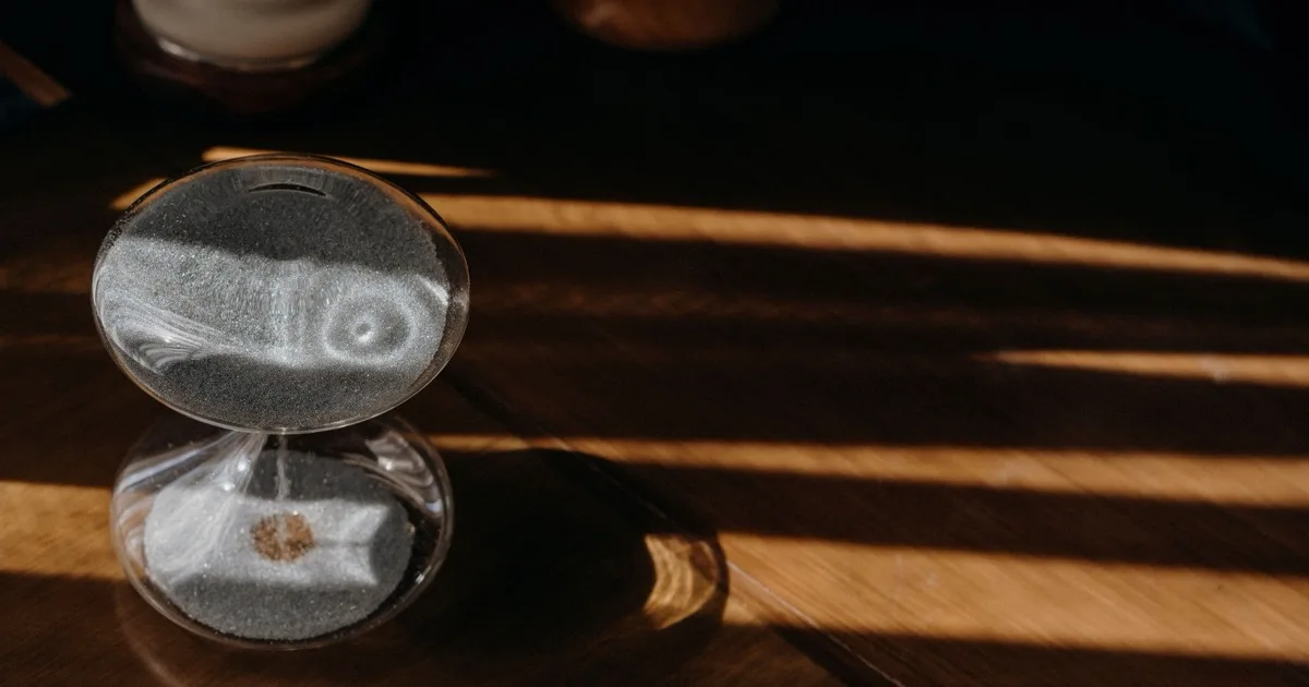 Reloj de arena sobre una mesa de madera, atravesado por franjas de luz de una persiana